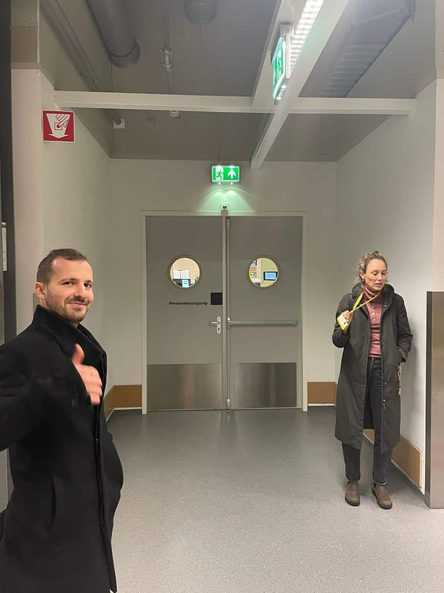 Two people standing in a hallway, one giving a thumbs-up, the other holding a drink.