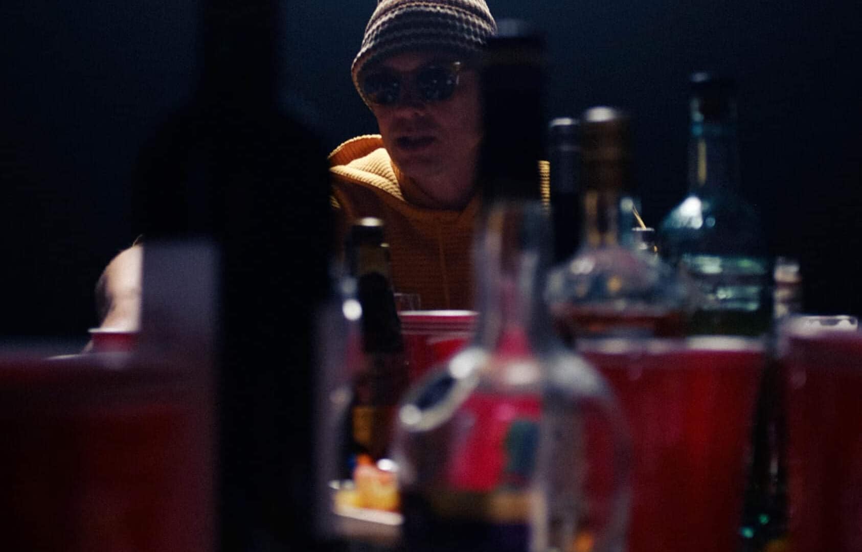 Person wearing a hat and sunglasses at a table with bottles and food, dim lighting.