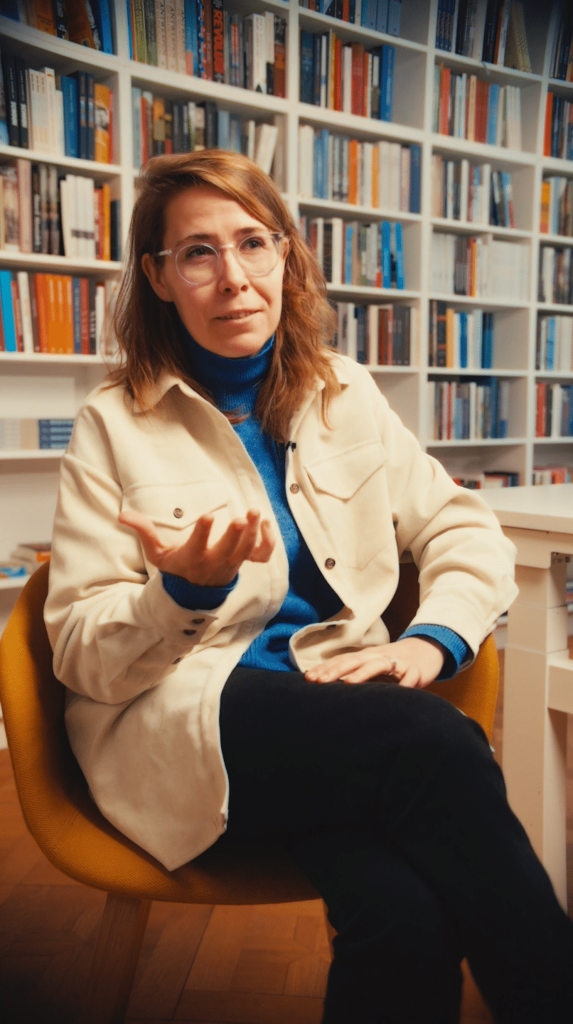Woman sitting in a library surrounded by bookshelves, engaging in conversation.